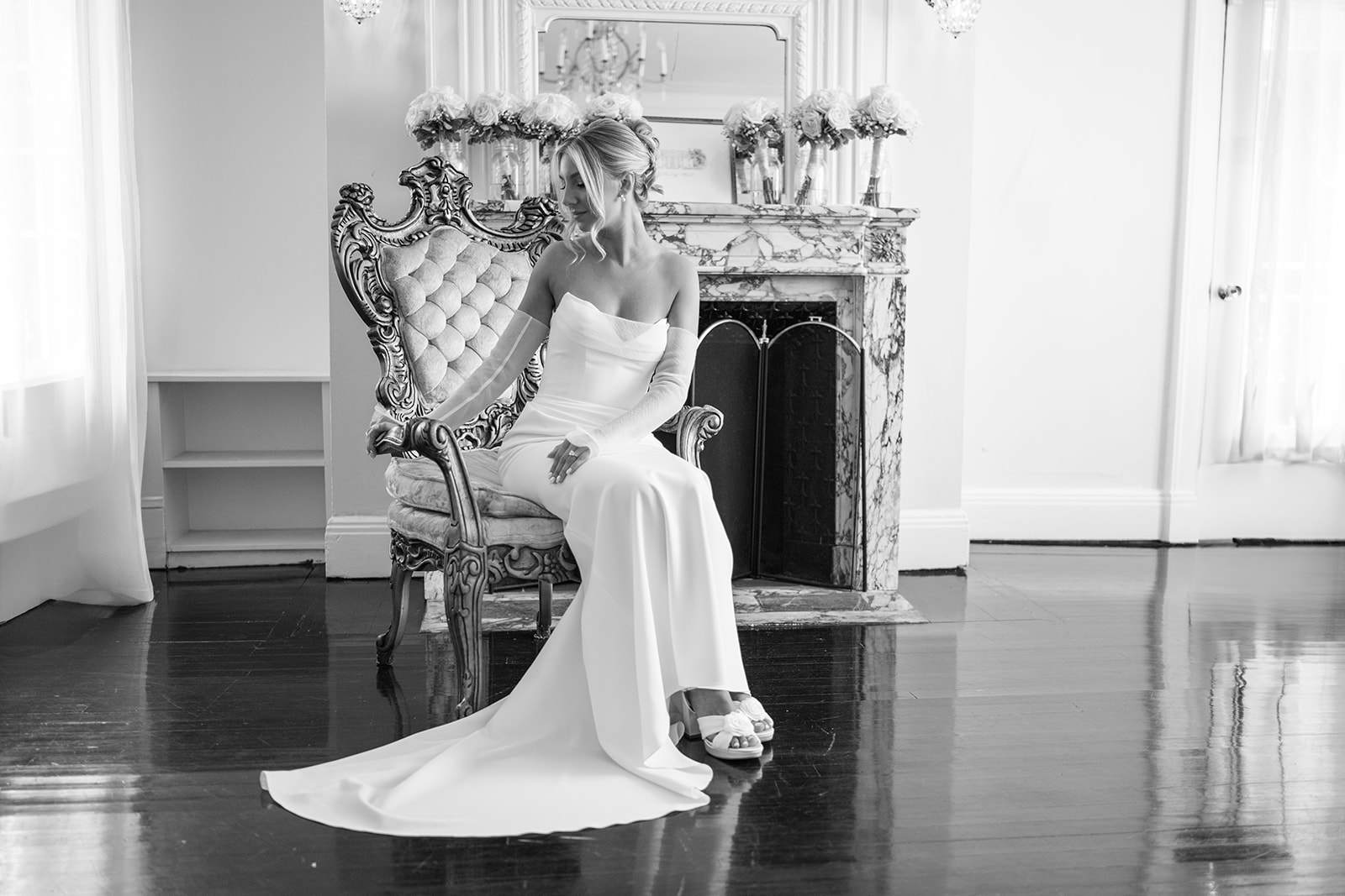 Black and white portrait of a bride sitting in a vintage chair beside a fireplace, looking down thoughtfully.